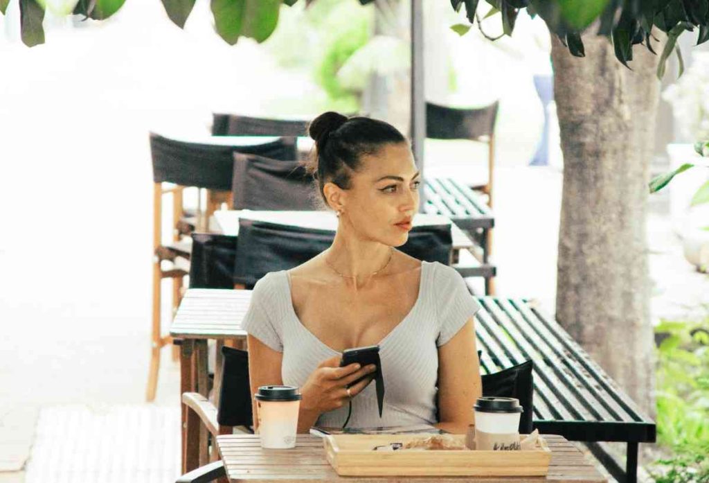 a woman holding a cell phone outside while eating and drinking coffee