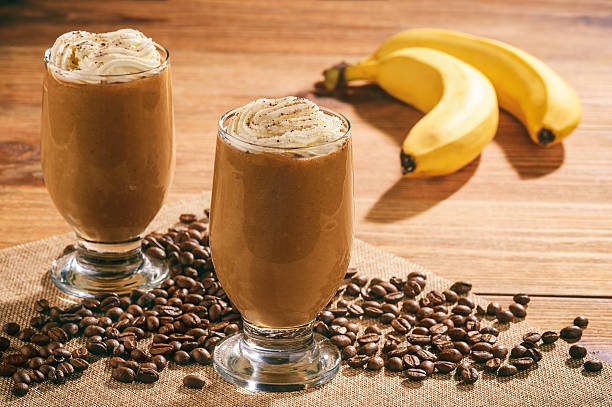 a coffee smoothie topped with whip cream with bananas and coffee beans on a wood table