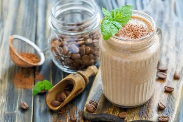 A creamy coffee smoothie served in a mason jar, topped with a sprig of mint and a dusting of cocoa or cinnamon. Surrounding the jar are whole coffee beans, a wooden scoop, and a glass jar filled with coffee beans, all set on a rustic wooden surface.