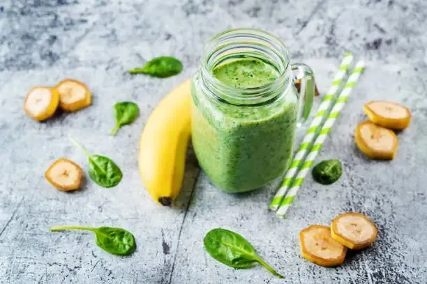 A vibrant Green Mocha coffee smoothie in a clear glass, placed in front of fresh spinach leaves, bananas, and a halved avocado. The ingredients suggest a healthy blend of spinach, banana, and avocado, lit with soft natural light.