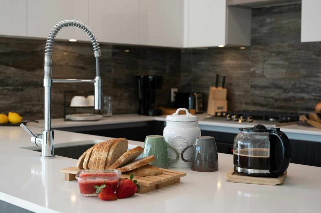 A modern kitchen countertop with a high-arc chrome faucet displays a cozy coffee setup, including a loaf of sliced multigrain bread, a small glass dish of strawberry jam with fresh strawberries, three ceramic mugs, a white coffee canister, and a half-full coffee pot on a wooden coaster. The background features a sleek dark backsplash, knife block, coffee maker, and minimalist decor, creating a stylish and inviting morning scene.