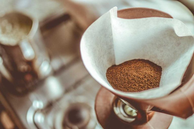 A close-up of ground coffee inside a white paper filter set in a ceramic pour-over coffee dripper, ready for brewing. The background is softly blurred, featuring a kettle and stove setup, highlighting a warm, homey coffee-making scene.