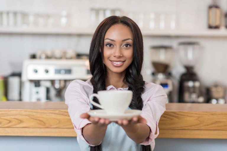 A smiling woman stands behind a café counter, holding a white cup and saucer toward the camera. The background features a coffee machine and shelves with cups.
