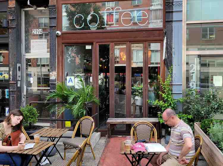 Two people sit at outdoor tables in front of a coffee shop with large glass windows and a sign reading coffee. Plants decorate the entrance, and the shops address, 2194, is visible on the door.
