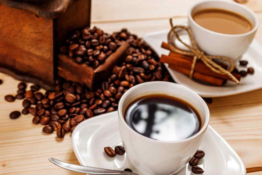 Two cups of coffee sit on white saucers with scattered coffee beans, a spoon, and cinnamon sticks. An open wooden grinder spills coffee beans onto a light wooden table in the background.