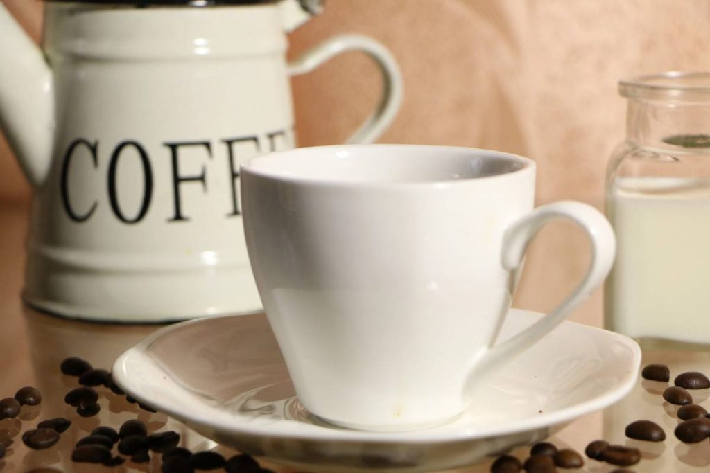 A white coffee cup and saucer are placed on a table surrounded by coffee beans. Behind the cup, there is a white pot labeled COFFEE and a glass jar filled with milk.