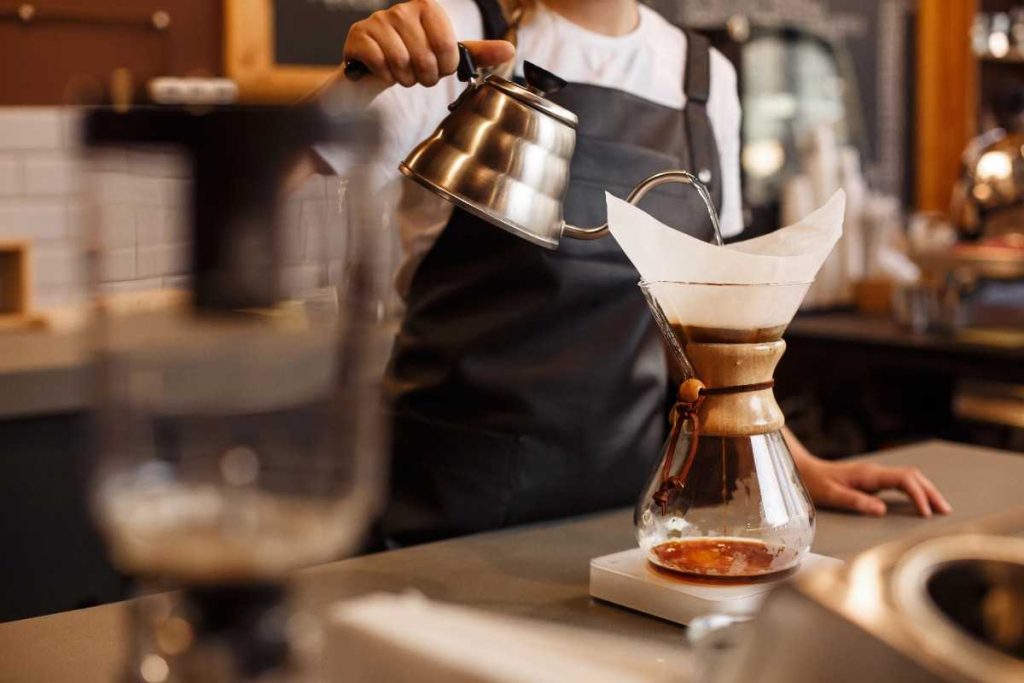 A person in a black apron pours hot water from a gooseneck kettle into a Chemex coffee maker with a paper filter, showcasing the pour-over method in a café setting—highlighting one option among French Press vs Pour Over vs Drip techniques.