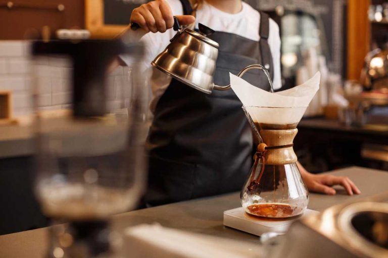 A barista wearing a dark apron pours hot water from a kettle into a Chemex coffee maker with a paper filter on a counter in a cozy café.