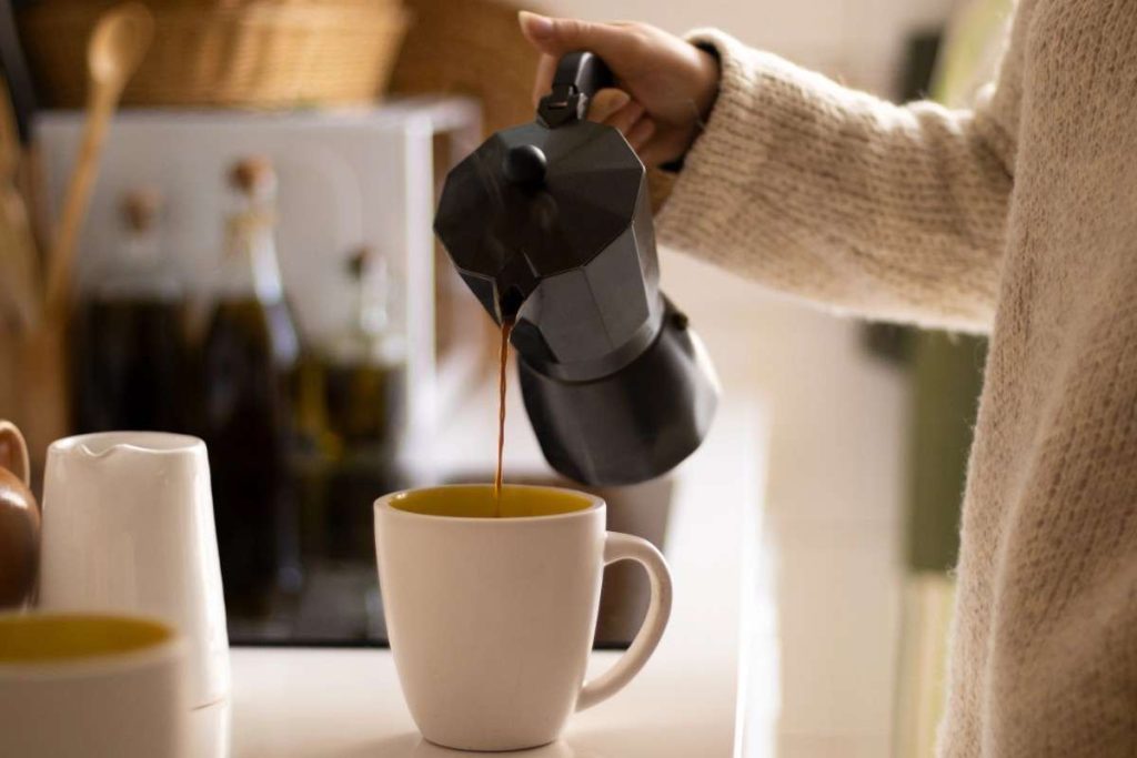 a woman pouring coffee from a black moka pot into a coffee cup