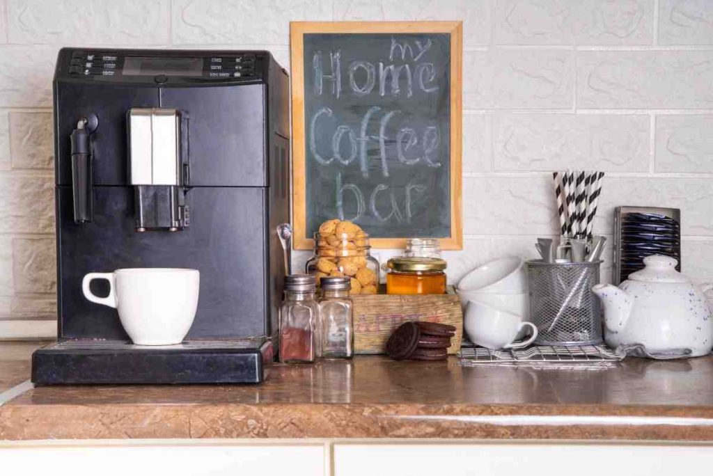 A kitchen counter displays a coffee machine with a white cup, jars of spices, cookies, honey, stacked cups, a teapot, and a chalkboard sign reading my home coffee bar in the background.