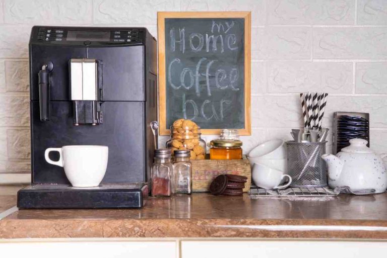 A kitchen counter displays a coffee machine with a white cup, jars of spices, cookies, honey, stacked cups, a teapot, and a chalkboard sign reading my home coffee bar in the background.