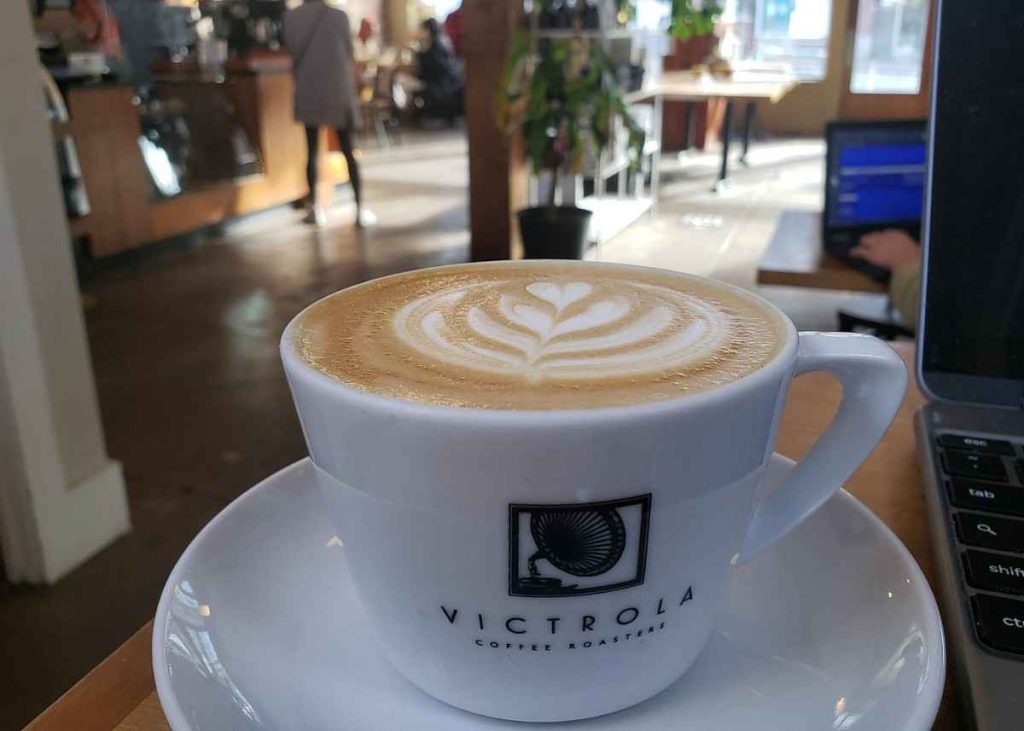 A cup of latte with heart-shaped foam art sits on a saucer at a café table, near a laptop. The café interior is bright, with patrons in the background and sunlight streaming through windows.