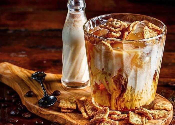 A glass filled with iced coffee, milk, and cereal pieces sits on a wooden board. Next to it is a small bottle of milk, a decorative spoon, and scattered cereal pieces and coffee beans.