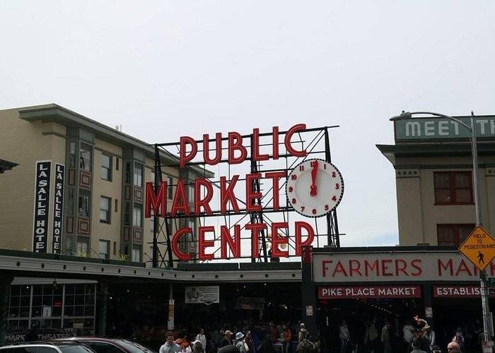 Large neon sign reading Public Market Center with a clock, above a crowded entrance to Pike Place Market in Seattle. Surrounding buildings include LaSalle Hotel and various market shops.