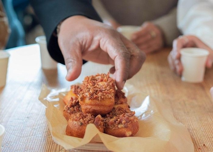 A close-up of a hand reaching to pick up a donut topped with crumbled bacon from a plate lined with parchment paper, with other people and small cups blurred in the background.