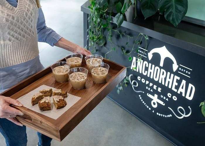 A person holds a wooden tray with four cups of iced coffee and several pieces of cake in front of an Anchorhead Coffee Co. sign and a counter with leafy plants.